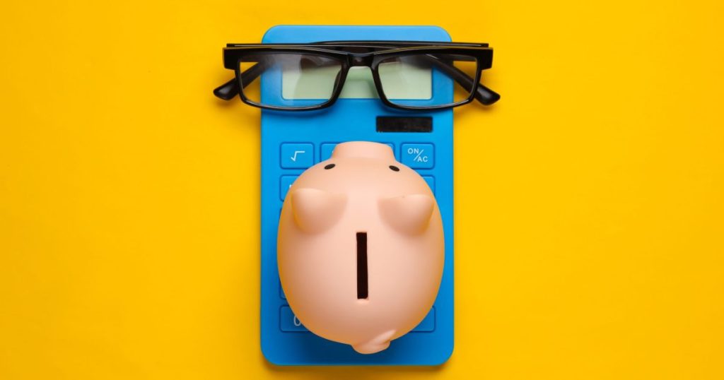 A piggy bank wearing reading glasses on top of a blue calculator, set against a yellow background